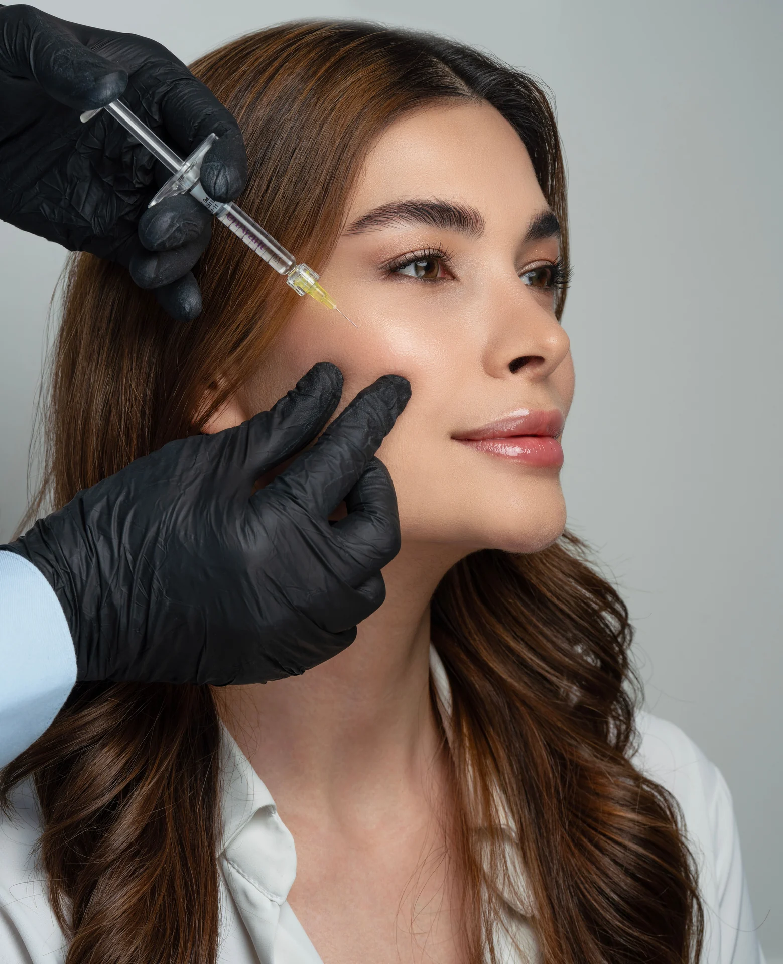 Imagem ilustrativa de aplicação facial com seringa próxima à bochecha; produto, paciente e profissional não identificados.